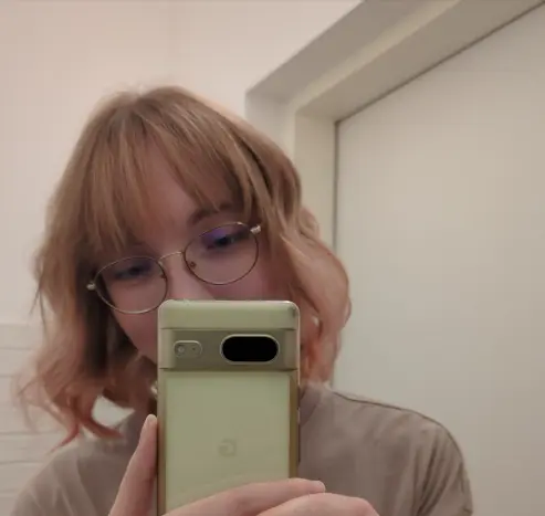 Mirror selfie in a small room. I have approximately chin-length hair which is pale brown (my natural hair color) at the top and gradually transitions to a warm rose at the bottom. It's wavy and I have slightly parted bangs going down to my eyebrows. I'm smirking behind my phone. The colors of the room and my shirt are creamy types of beige which goes nicely with my hair.