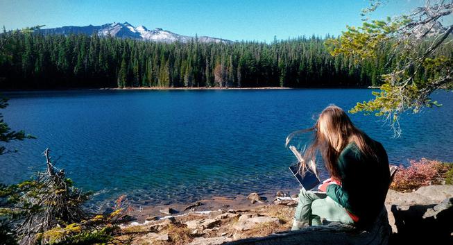 a photograph of this one sitting on a lake shore. a mountain big slowly sloping with a bit of snow is visible to the left across the lake, and to the right there is a barren stone peak partially obscured by a tree branch. I am seated on a log wearing jeans and two layers of jacket with a laptop on my lap and my hair blowing in the wind. the lake is a very deep blue while still being clear enough to see beneath it in shallow water. the shore on my side of the lake is rocky, which there are a few sandy beach protrusions on the far shore. between the far shore and the mountains, there is a thick evergreen forest