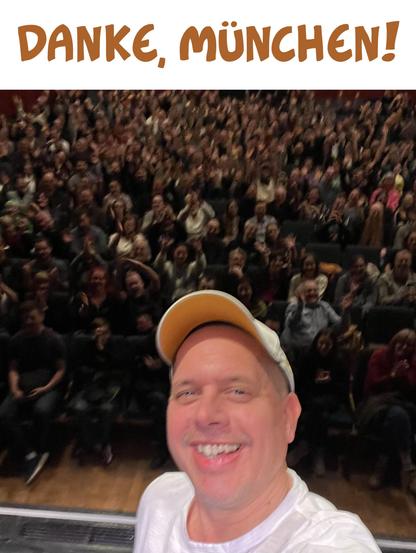 Ein Foto von Witzbildmaler Ralph Ruthe. Er steht auf der Bühne, grinst. Im Hintergrund sieht man einen mit mehreren hundert Menschen gefüllten Saal. Darüber steht "Danke, München!"