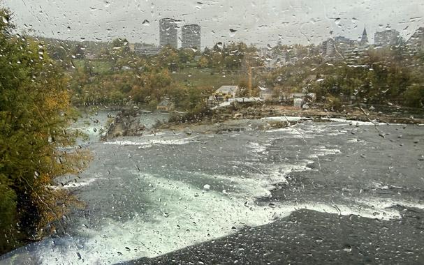 Blick aus dem Zugfenster auf den Rheinfall von der Bahnstrecke Schaffhausen – Winterthur bei regnerischem Wetter. Der Blick ist erschwert, da auf dem Fenster viele Regentropfen sind.