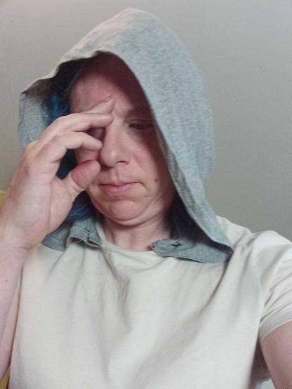 A person facing down and to the left of the camera, wearing a beige t-shirt with grey hood. Their hand is over their face.
