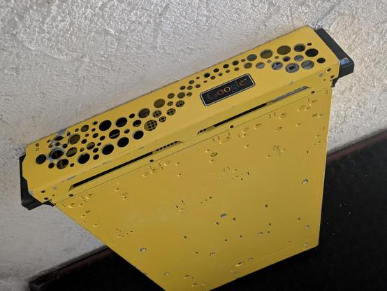 a cute brigh yellow google server leaning against a wall, on the front it appears to have a fan exhaust of a wave-like plane of holes of variable size